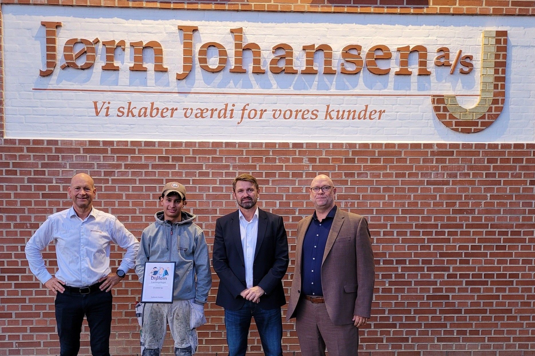 Jørn Johansen (t.v.) står stolt ved siden af sin murerlærling Sebastian Halloumi, som igen står ved siden af Anders Møller og Kasper Bæk fra Sydbank. Bagved syner logoet, som Halloumi har været med til at lave. Foto: Presse